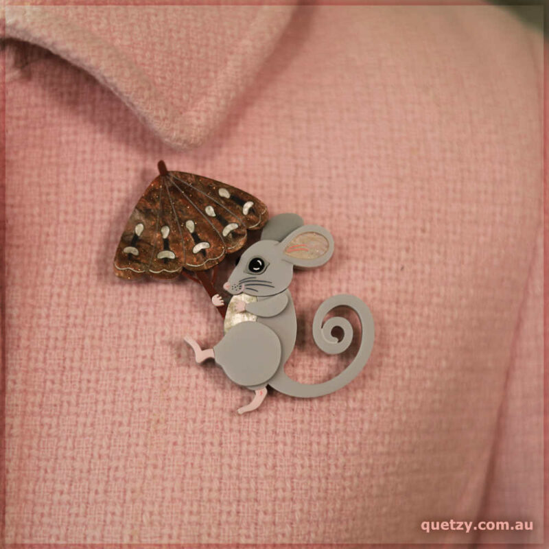 An acrylic brooch of a parading Mountain Pygmy Possum shaded by a Bogong Moth umbrella. Displayed on a pink coat.